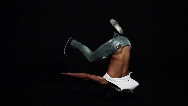 Young athlete active male break dancer dressed in white shirt and blue tight jeans, wearing bike helmet is actively spinning on his head on black background