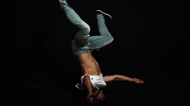 Young athlete attractive male break dancer dressed in white shirt and blue tight jeans, wearing bike helmet is actively spinning on his head on black background