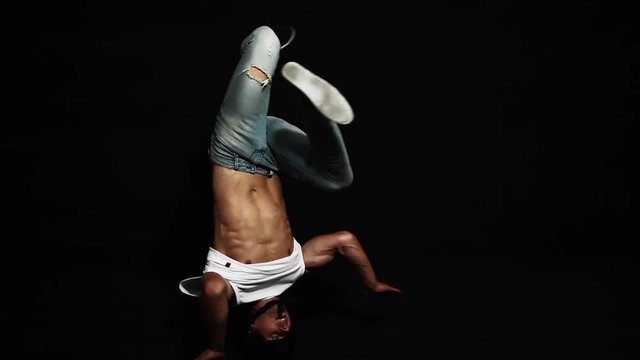 Young athlete attractive male break dancer dressed in white shirt and blue tight jeans, wearing protective bike helmet is actively spinning on his head on black background