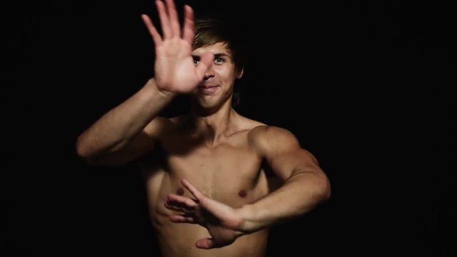 Young athletic attractive blue eyed male with naked torso and having modern haircut is smiling and dynamically waving his hands in front of camera on black matte background.