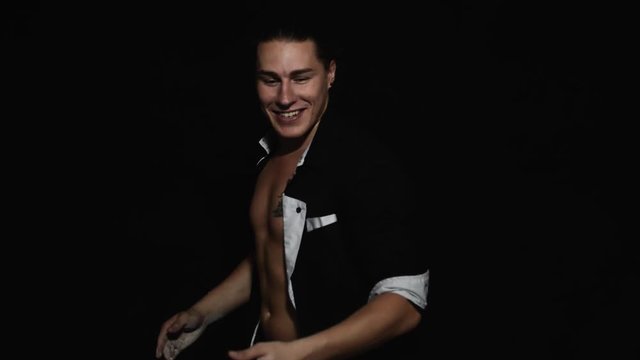 Handsome athletic young brown eyed guy in unbuttoned shirt with tattoo on chest is smiling and dynamically clapping with hands covered by white powder in front of camera on black matte background.