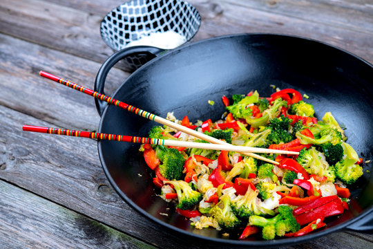 wok with vegetables, bowl of rice and chopsticks on wooden groun