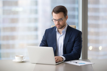 Serious millennial businessman working at laptop typing business letter drinking morning coffee in...