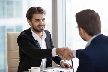 Smiling millennial male CEO shaking hand of business partner during office meeting, satisfied businessman handshaking colleague at negotiations, boss greeting or thanking for briefing. Human resources