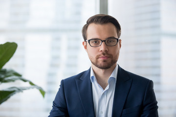 Portrait of serious millennial businessman wearing glasses looking at camera, headshot of concentrated confident male worker or director posing in modern office, making photo or picture near window