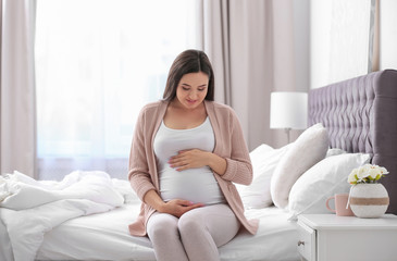 Young beautiful pregnant woman sitting on bed and touching her belly at home