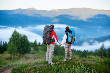 Obraz premium Rear view man and woman with backpacks holding hands on top of a hill with a view of the mountains in a light haze and a path to them. Concept of active outdoor hiking lifestyle