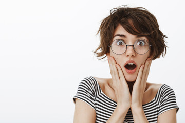 Impressed and shocked charming female with stylish short haircut in round trendy glasses, gasping and dropping jaw, holding palms on cheeks, being stunned with news or rumors spread across office