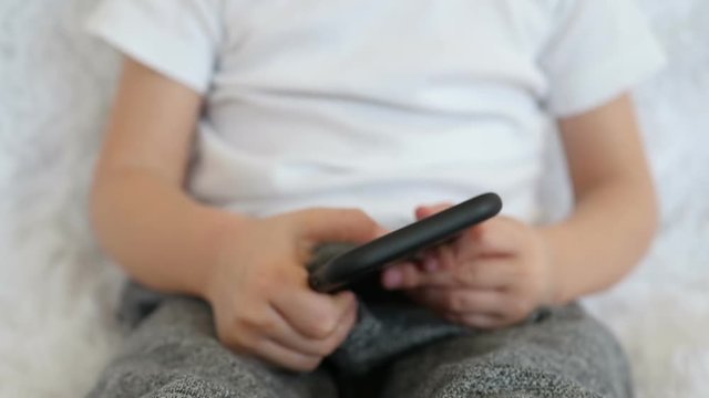 The child is playing with a smartphone. Move the camera to your feet, phone and face of the child, close-up