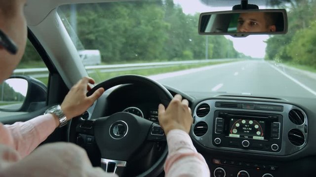 Reflection in rear view mirror of confident handsome man driving car on empty asphalt road. Male driver driving auto on freeway during business travel. View from inside of vehicle interior.