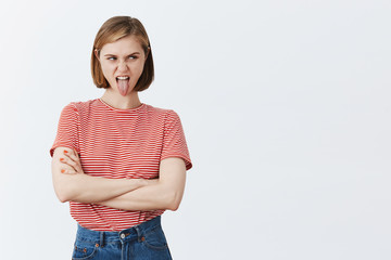 Moody and reckless girlfriend showing character unwilling to go on mountain trip with boyfriend, holding hands crossed on chest in offended pose, showing tongue and looking right from under forehead