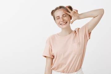 Joyful good-looking young girl with buns hairstyle and freckles enjoying carefree life, smiling broadly with closed eyes, showing peace gesture near face, being happy and enthusiastic over gray wall