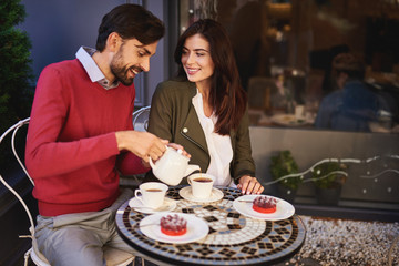 Love and care. Portrait of beautiful young couple sitting at the table in outdoor cafe. Gentleman using teapot while lady looking at him with smile