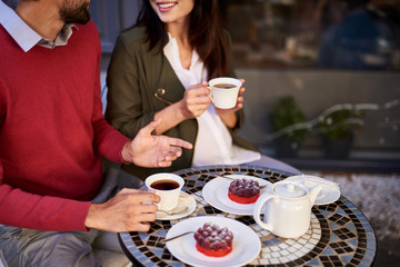 Fototapeta na wymiar Perfect date. Cropped smiling lady and bearded man sitting at the table with cups of tea, raspberry cakes on plates and teapot