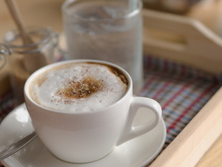 Hot cappuccino mug with white foam brown sugar topping in white cup drinking water in glass closeup