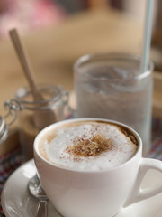 Hot cappuccino mug with white foam brown sugar topping in white cup drinking water in glass closeup