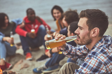 On same wave. Portrait of young guy drinking beer from glass bottle with his friends on background