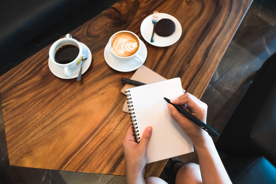Top view image of a woman's hands holding and writing down on a white blank notebook with coffee cups on wooden table in cafe