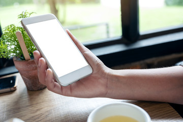Mockup image of a woman's hands holding a white mobile phone with blank screen with coffee cup and tea on table in cafe