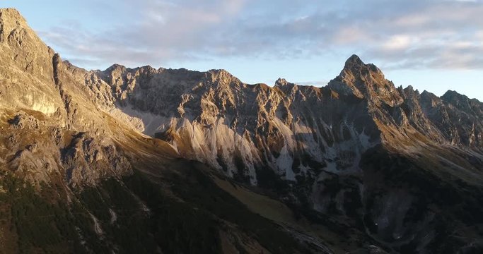 Cinematic view of alps mountain ridge and summit peak Zimba, Austria during sunrise. Drone filming moving sideways filming rocky mountains and green valley with hut cabin.