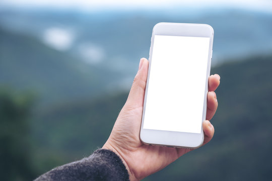 Mockup image of a hand holding and showing white smart phone with blank desktop screen in outdoor with blur green mountains background