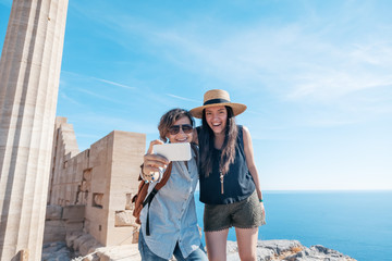 Fototapeta premium Two young beautiful happy woman female travel girlfriends doing selfie on the background of the sea and ancient ruins in Greece