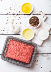 Tray with raw minced homemade beef meat with spices and herbs. Top view. On top of wooden kitchen table background. With pepper salt and tomatoes.