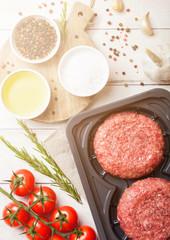 Plastic tray with raw minced homemade beef burgers with spices and herbs. Top view. On top of wooden kitchen table background with tomatoes salt and pepper.