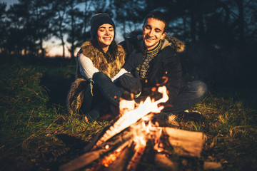 Pretty couple relaxing near bonfire in the forest at evening time