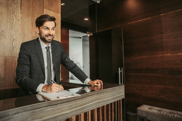 Portrait of cheerful bearded man writing on paper while locating at counter during job in modern hotel indoor