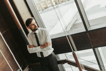 Portrait of concentrated bearded man with crossing hands standing near window and looking away during break at job