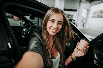 Fototapeta na wymiar Portrait of happy female taking selfie while drinking mug of appetizing liquid during rest in contemporary vehicle