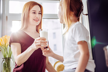 Glass of milk. Cheerful emotional young woman kindly looking at her little daughter and smiling...