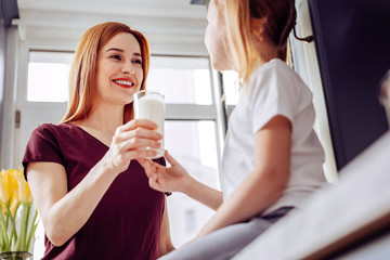 Healthy lifestyle. Positive enthusiastic young woman taking care of the health of her little daughter and giving her a cup of milk