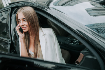 Portrait of happy pretty female telling on mobile while looking from opened window of contemporary car. Satisfied lady during communication on gadget in vehicle concept