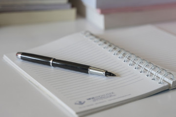 Close up of notebook with ink-pen and eyeglasses put on white table with blur focus of books as a background. Educational concept.
