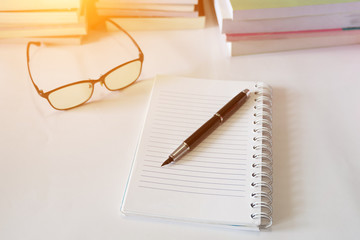 Close up of notebook with ink-pen and eyeglasses put on white table with blur focus of books as a background. Educational concept.