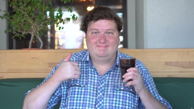 Fat happy man holding a glass of drink cola and showing thumb up gesture, sitting in cafe. Beverage