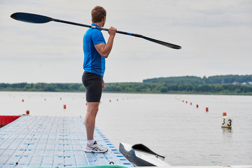 Young athlete in sportswear standing with paddle on the pier and looking at view of calm lake