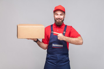Young cheerful logistic delivery man with beard in blue uniform and red t-shirt standing, holding...