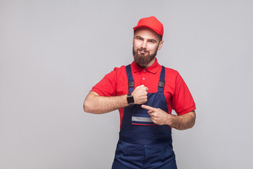 We do ontime. Young confident handyman with beard in blue overall and red t-shirt standing and showing time on his wrist watch with smile. Grey background, indoor, studio shot, isolated