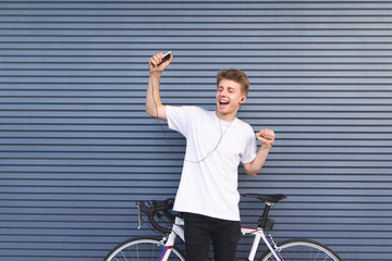 Happy young man standing by a white bicycle, listening to music in headphones with a smartphone in...