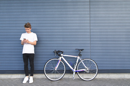 Young man in a white shirt standing near the road bike and uses the smartphone. Happy student standing by a bicycle on the background of the wall.