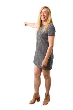 Middle age blonde woman with grey dress pointing back with the index finger presenting a product on isolated white background