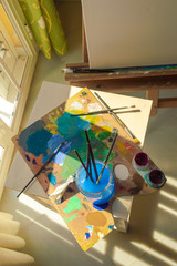 Materials to paint with sunbeams of a window: jar with water to clean the brushes, blank canvas on easel and used palette full of colorful paint.