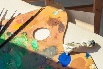 Materials to paint: blank canvas, palette full of colorful paint, brushes and oil paint tube with sunbeams entering through a window.
