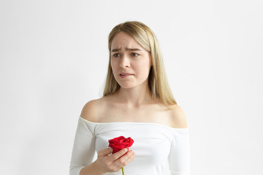 Picture of frustrated beautiful young lady posing in studio with one red rose from her lover, being worried and upset, biting lips, doesn't know how to say husband that she loves other man