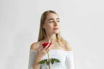 Romantic young Caucasian female wearing open shoulders long sleeved dress holding one red rose from guy on first date, looking sideways with dreamy mysterious smile. Love, passion and romance