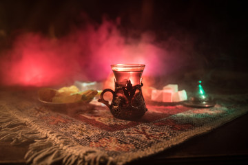 Arabian tea in glass with eastern snacks on a carpet on dark background with lights and smoke. Eastern tea concept. Empty space.