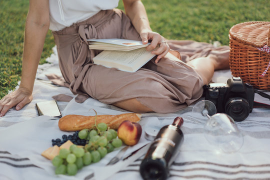 Cropped horizontal picture of girls legs having picnic on a grass in the Parc with blank bottle of wine and appetizers and a photo camera and reading a book. Blogger taking pictures of a picnic
